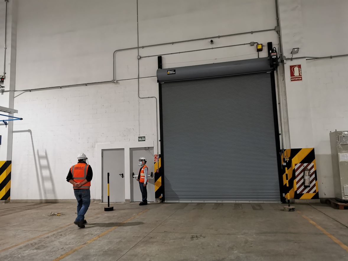 Puerta enrollable cortafuego de gran tamaño instalada en entorno industrial, diseñada para activarse automáticamente en caso de incendio, formando una barrera que sectoriza el fuego, con operarios que evidencian la escala de la instalación.