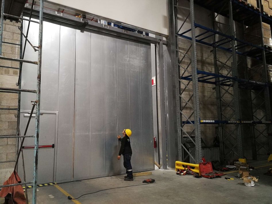 Puerta corrediza cortafuego en proceso de instalación en exterior de almacén, con paneles tipo sándwich unidos mediante sistema macho-hembra y núcleo de lana de roca que aporta resistencia al fuego y aislamiento térmico, con operario en escena.