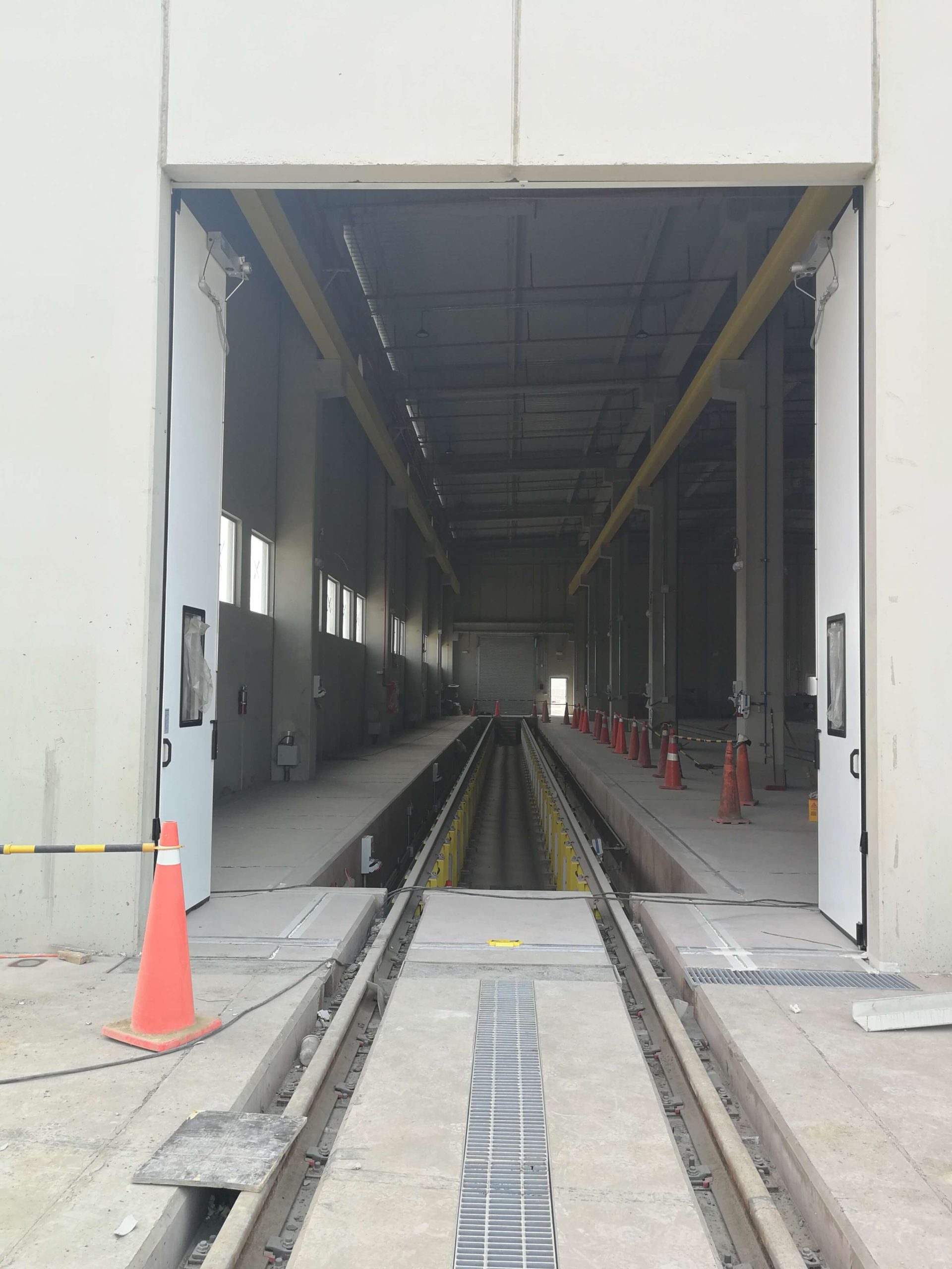 Puerta abatible industrial en color blanco abierta en patio taller del tren eléctrico, con sistema de plegado lateral que maximiza el espacio de apertura, diseño sin riel inferior para paso libre y opción de visores que mejoran la visibilidad en operación.