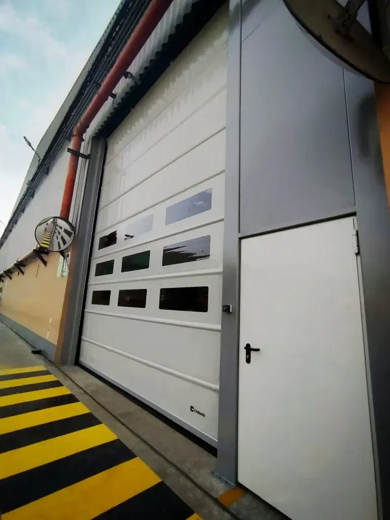 Puerta rápida de PVC FastPack Folding en color blanco instalada en entorno logístico exterior, con sistema de izaje por correas y refuerzos transversales que aportan estabilidad ante el viento en vanos de gran dimensión.