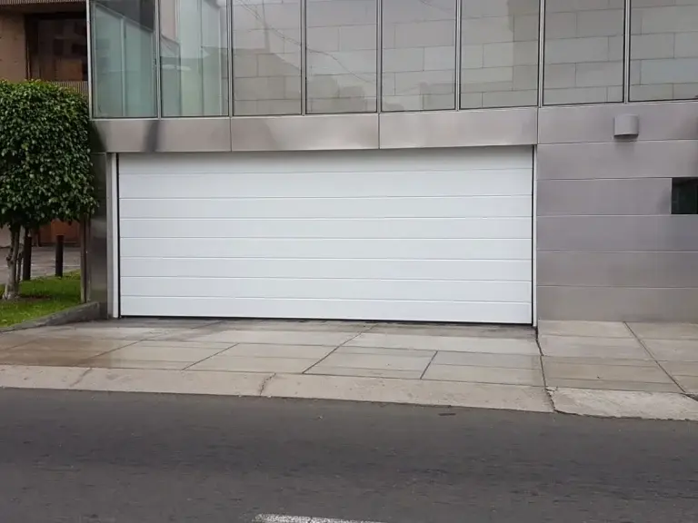 Puerta seccional de garaje en color blanco instalada en edificio residencial, con apertura vertical que permite un mejor aprovechamiento del espacio, acabado de calidad y diseño integrado a la arquitectura del entorno.
