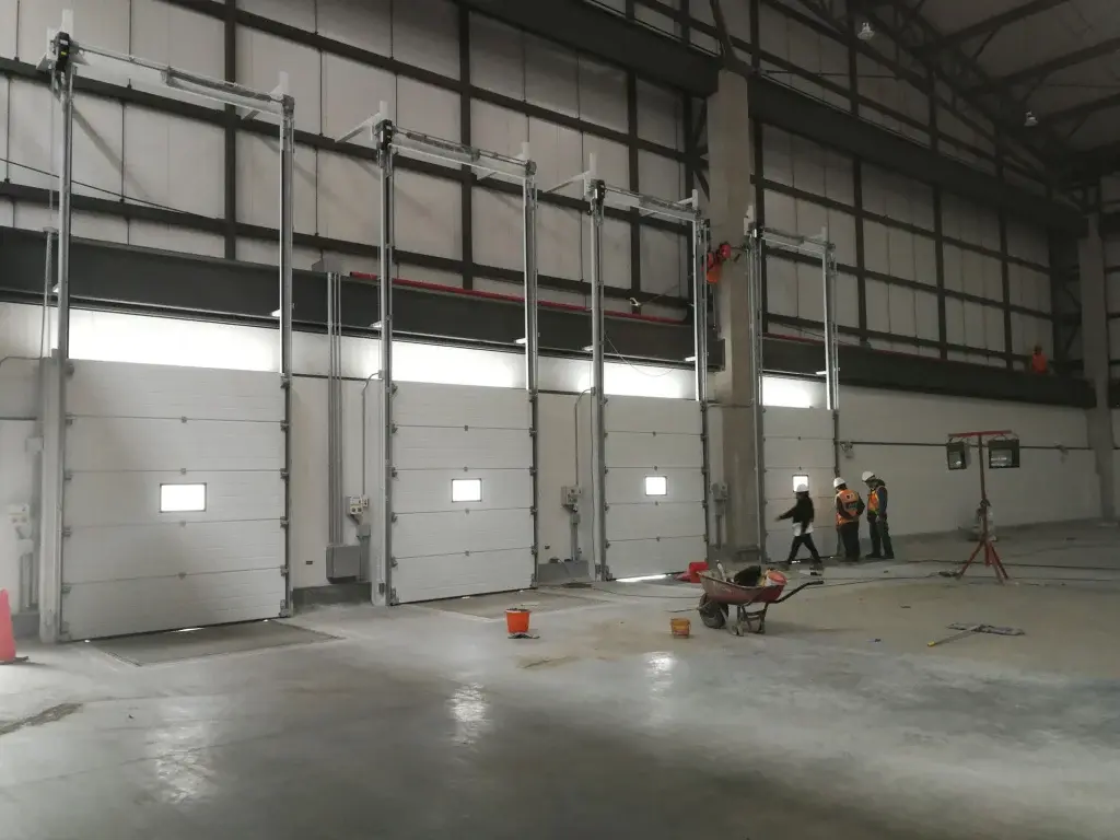 Puertas seccionales industriales en color blanco dentro de almacén, con operarios en zona de trabajo, equipadas con paneles modulares tipo sándwich que brindan aislamiento y cierre eficiente.