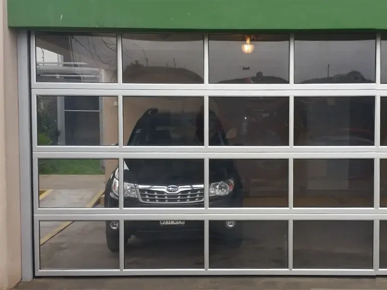Puerta seccional polivista de garaje instalada en vivienda, fabricada con perfiles de aluminio y paneles de policarbonato que permiten el ingreso de luz natural y visibilidad hacia el interior, brindando seguridad en el acceso vehicular.